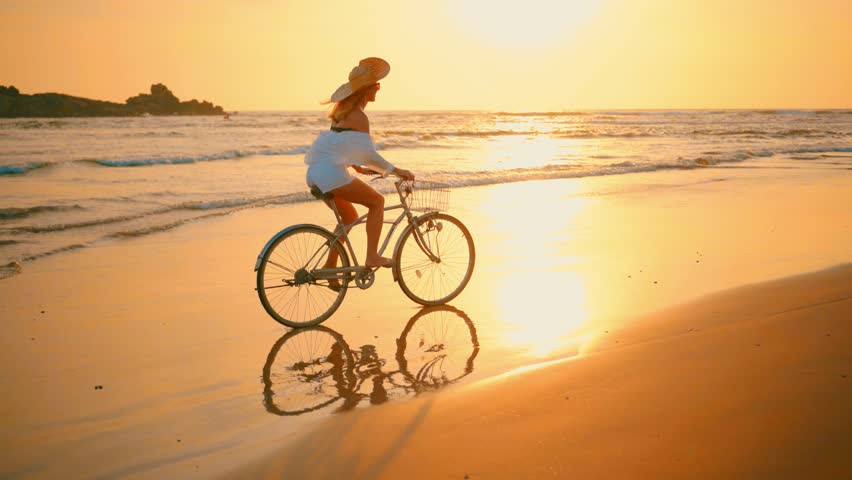 Woman in white and hat mooving on her bicycle along beach sand at summer time under burning sunset sky, lifestyle carefree.