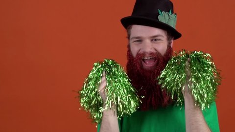 Young man celebrating saint patrick's day isolated on orange dancing with pom poms