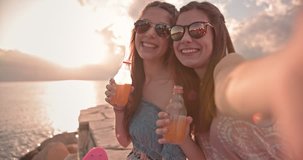 Teenage best friends taking selfies on smartphone, relaxing by the sea and drinking fizzy drinks