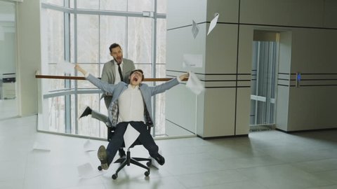 Slow motion of Two crazy businessmen riding office chair and throwing papers up while having fun in lobby of modern business center