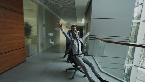 Dolly shot of Two crazy businessmen riding office chair and throwing papers up while having fun in lobby of modern business center
