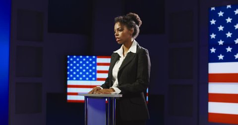 Confident African-American woman holding press conference while standing alone on podium with background of American flag on screen.