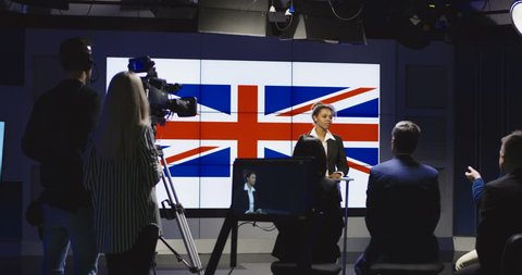 Production team giving instructions to a female news anchor as she stands in front of the British flag with a cameraman filming to the left. 4K shot on Red cinema camera.