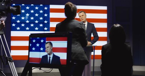 Official press conference of American representative politician on stage against display with American flag giving speech to audience in semilit studio and answers the questions.