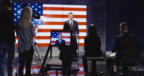 Official press conference of American representative politician on stage against display with American flag giving speech to audience in semilit studio. 4K shot on Red cinema camera.