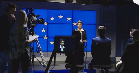 Serious African-American representative of European Union answering questions of journalists on news conference in semilit studio with EU flag on screen. 4K shot on Red cinema camera.