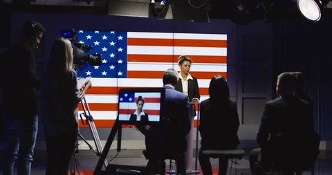 Production team filming a show host interviewing a group of people in a broadcast studio with an American flag backdrop