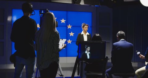 Female news anchor reading the news in front of a European flag with a production team and camera man in the studio