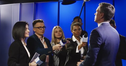 Big group of multiracial journalists with microphones and other technological equipment having interview with middle-aged politician in underlit studio.