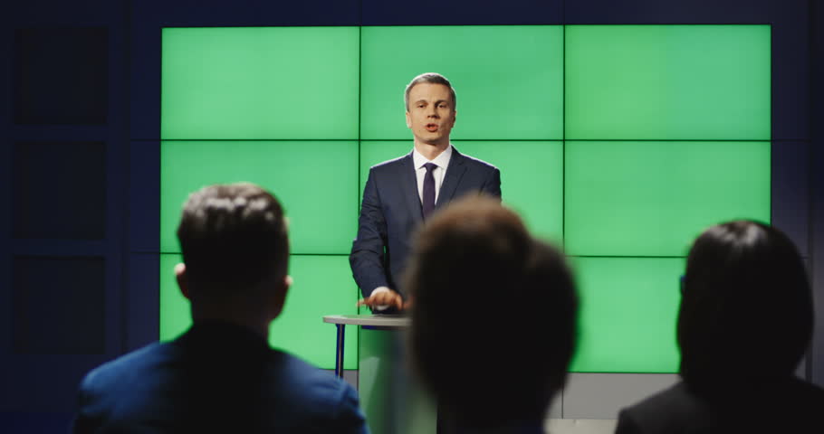 View from behind a production team in a studio sitting listening to a presenter talking in front of a green screen then breaking into applause clapping.