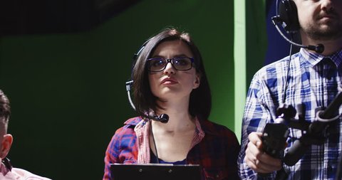 Female producer or prompt wearing a headset and holding a clipboard alongside a cameraman in a production studio panning to male colleague on the left of the screen.