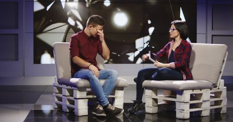 Young confident and relaxed man and woman taking participation in television show sitting in modern illuminated studio and having talk.