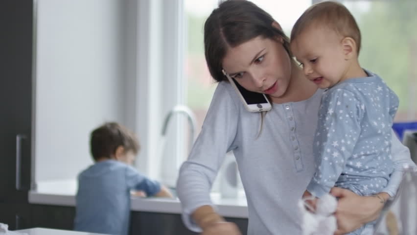 Mother cleaning kitchen table while holding baby and talking on the phone, her son doing dishwashing in the background Royalty-Free Stock Footage #1007751085