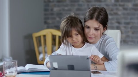 Mother using tablet computer for remote video communication and helping her daughter with homework, her son is busy in the background