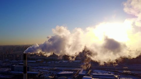 White exhaust, flue gas from central heating station chimney. Aerial shot in bright sun, camera fly towards curling clouds of smoke and water vapour. Dark shaded cityscape, sleeping quarters buildings