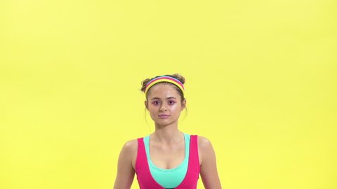 Retro portrait closeup of young woman in colorful sportswear lifting weights while doing aerobics in gym, isolated over yellow background slow motion retro style