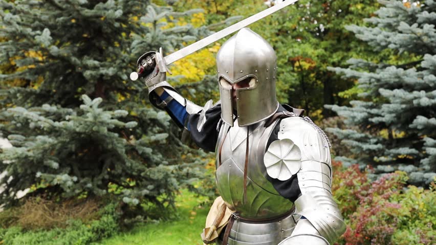 Close-up of man in medieval iron armor threatens with sword in hand standing on green grass, slow motion