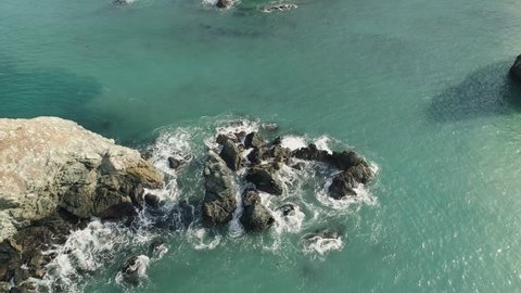 Aerial shot looking down and revolving around rocks in the ocean. Swirling waves with turquoise water and big rocks.