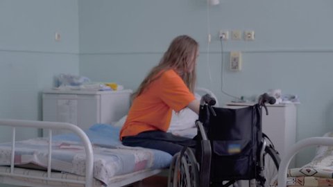 Young guy, a disabled person with a lesion of the spinal cord and legs, multiple sclerosis, without assistance, is transplanted from a hospital bed to a wheelchair. 