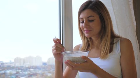 A young pretty brunette woman eats cereal with milk from a bowl. Standing at the window and looks outside. Healthy lifestyle and nutrition. Good mood and pleasure. White linen, 4k, 3840x2160.