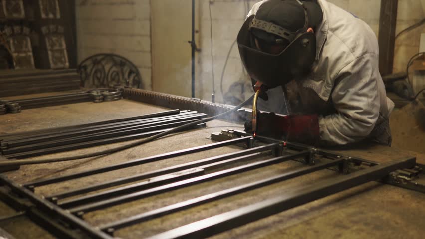 Welder is welding parts of steelwork on a table. He is using powerful industrial welding machine, in workshop of forged products