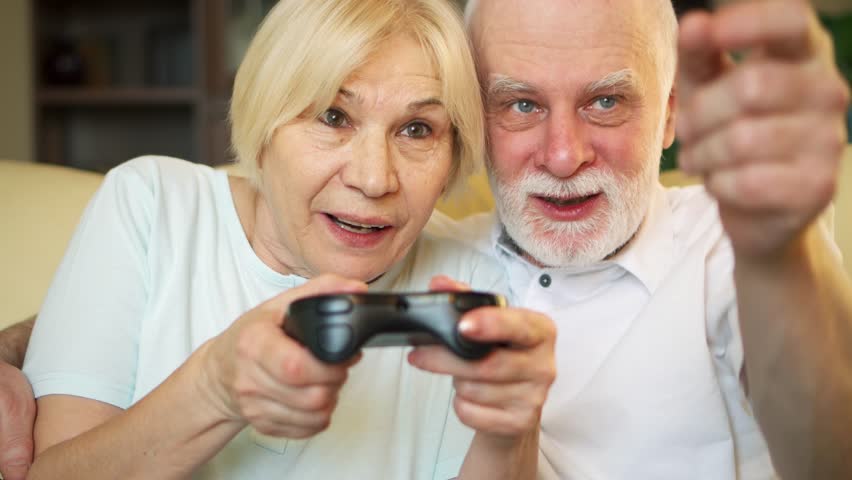 Cheerful senior couple gamers playing video games at home. Addicted seniorplayers with remote controller of game console. Cybersport for active modern elderly people | Shutterstock HD Video #1007808682