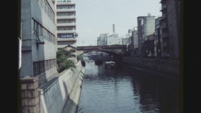 TOKYO, JAPAN, MARCH 1978. Ginza City Impression Of River Channel Between Buildings With Freight Boats And A Train Passing Over A Bridge