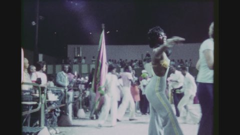 RIO DE JANEIRO, BRAZIL, MARCH 1976. Young Brazilian Woman Flagbearer Dancing Samba Amongst Other Skillful Dancers In A Public Open Air Terrace Club.