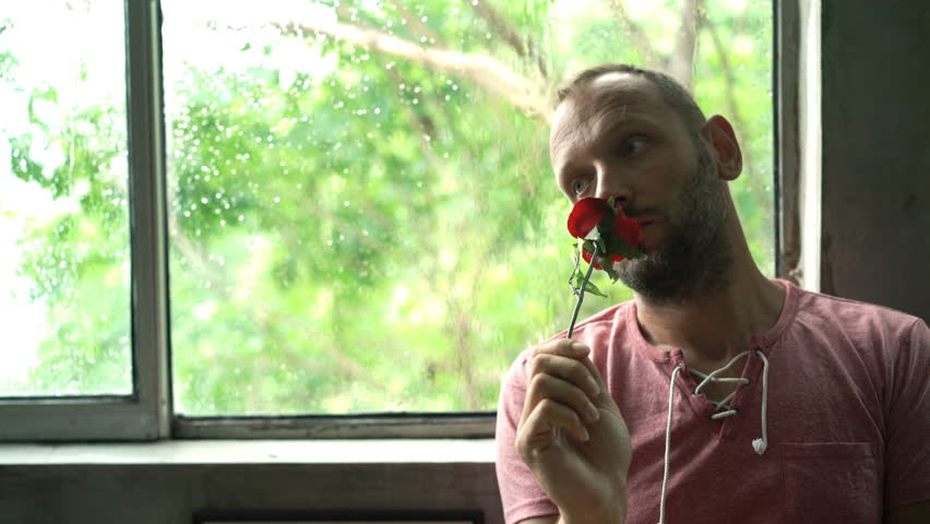 Sad, pensive man holding red rose sitting by window during rainy day 
 | Shutterstock HD Video #1007826688