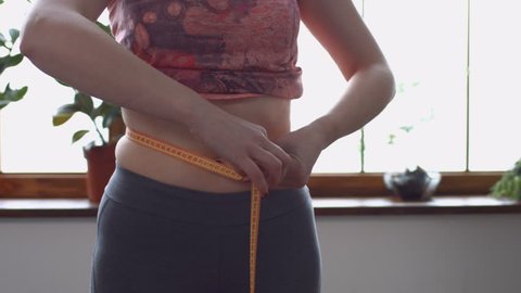 Midsection of adult woman measuring her waistline with measure tape while standing in domestic room. Closeup. Positive woman checking success of weight loss program with measuring tape at home.