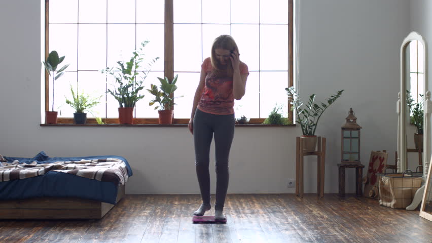 Happy attractive redhead woman celebrating and cheering a weight loss goal achievement while standing on weighing scales in domestic room. Positive woman jumping and clapping hands after weight loss.
