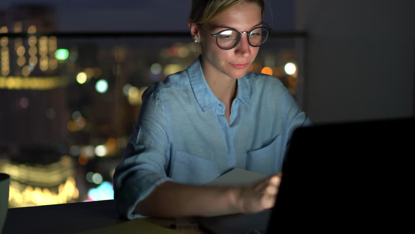Smiling attractive female in eye glasses with reflection of screen lights reading news on smartphone in social network and using laptop computer while sitting at night with darkness in home interior Royalty-Free Stock Footage #1007840560