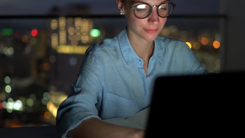Smiling attractive female in eye glasses with reflection of screen lights reading news on smartphone in social network and using laptop computer while sitting at night with darkness in home interior
