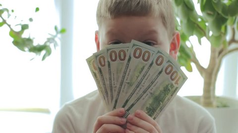 Happy boy in the background of money trees keeps a hundred dollar bills and hides behind them. The concept of savings and investment from an early age. 