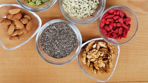Superfood seeds and nuts and berries, chia, almond, walnut, hemp, pumpkin, goji in glass bowl over wooden table, slow motion