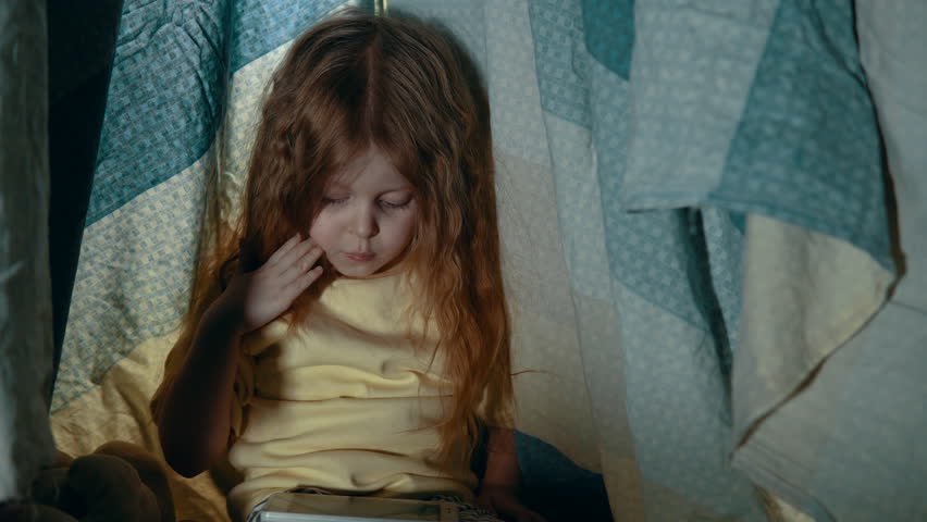 A little girl in pajamas uses the phone while sitting in a tepee tent in the playroom in the evening.