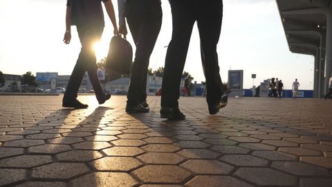 Feet of three businessmen walking near airport with sun flare at background. Business men go to terminal together. Confident guys being on his way to work trip. Colleagues going outdoor. Slow motion