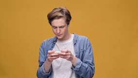 Young concentrated guy standing isolated over yellow background play games by mobile phone