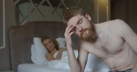 Close-up portrait of the depressed ginger head man sitting on the bed at the blurred background of his blonde wife lying on it.
