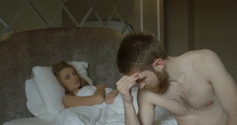 Depressed ginger head man is worrying about problems in bed at the blurred background of his disappointed blonde wife turning back.
