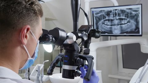 dentist in medical mask adjusts binocular microscope to work, roentgen of jaw on screen on background วิดีโอสต็อก