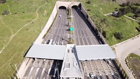 Highway traffic entering and exiting a Toll collection stop, leading to a double tunnel surrounded by green landscape - Aerial footage