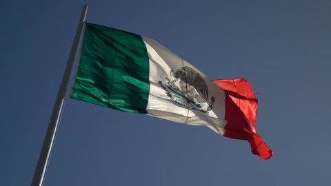 Facing right, large slow motion of back lit Mexican flag waving in the wind.