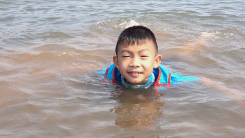 Swimming in summer concept. Asian boy slow motion swim float on water play in lake funny. He wearing life jacket for safety. View see mountain and sky background beauty. This Pattaya lake in Thailand