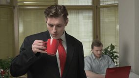 Young guy in a suit is standing in the office, smiling and drinking coffee, tea. The guy is working on the laptop in the background 60 fps