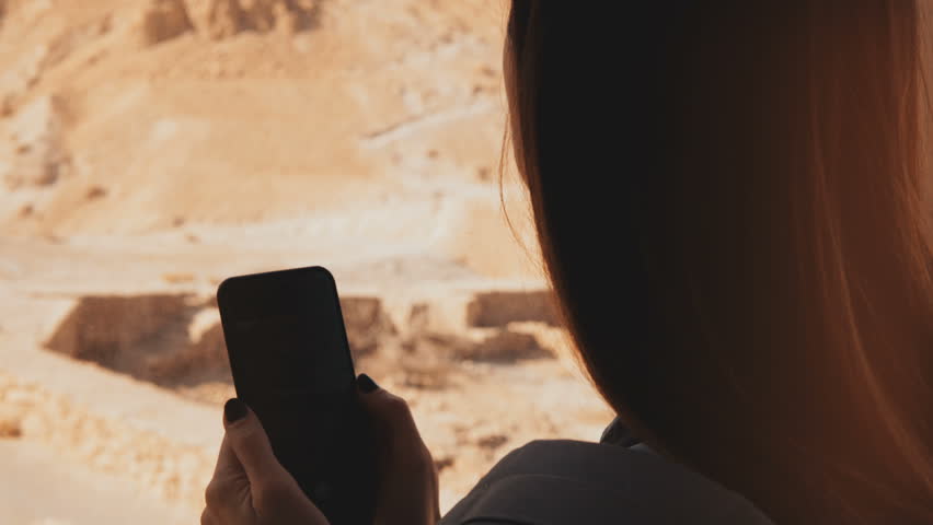 Girl using smartphone in aerial railway cabin. Woman takes phone photos in sunny desert ropeway car. Masada, Israel. 4K