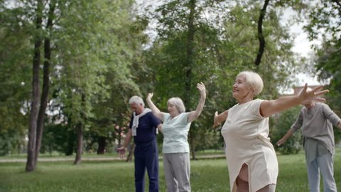 Tracking shot of group of elderly people doing warming up exercises, stretching and breathing deeply in and out in park