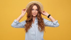 Cheerful ginger woman in denim shirt and hat moving and looking at the camera over yellow background