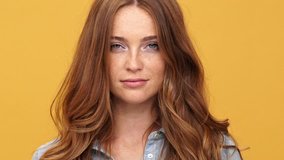 Close up of Smiling ginger woman in denim shirt playing with her hair and looking at the camera over yellow background