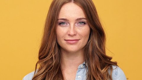 Close up of Smiling ginger woman in denim shirt winks and looking at camera over yellow background